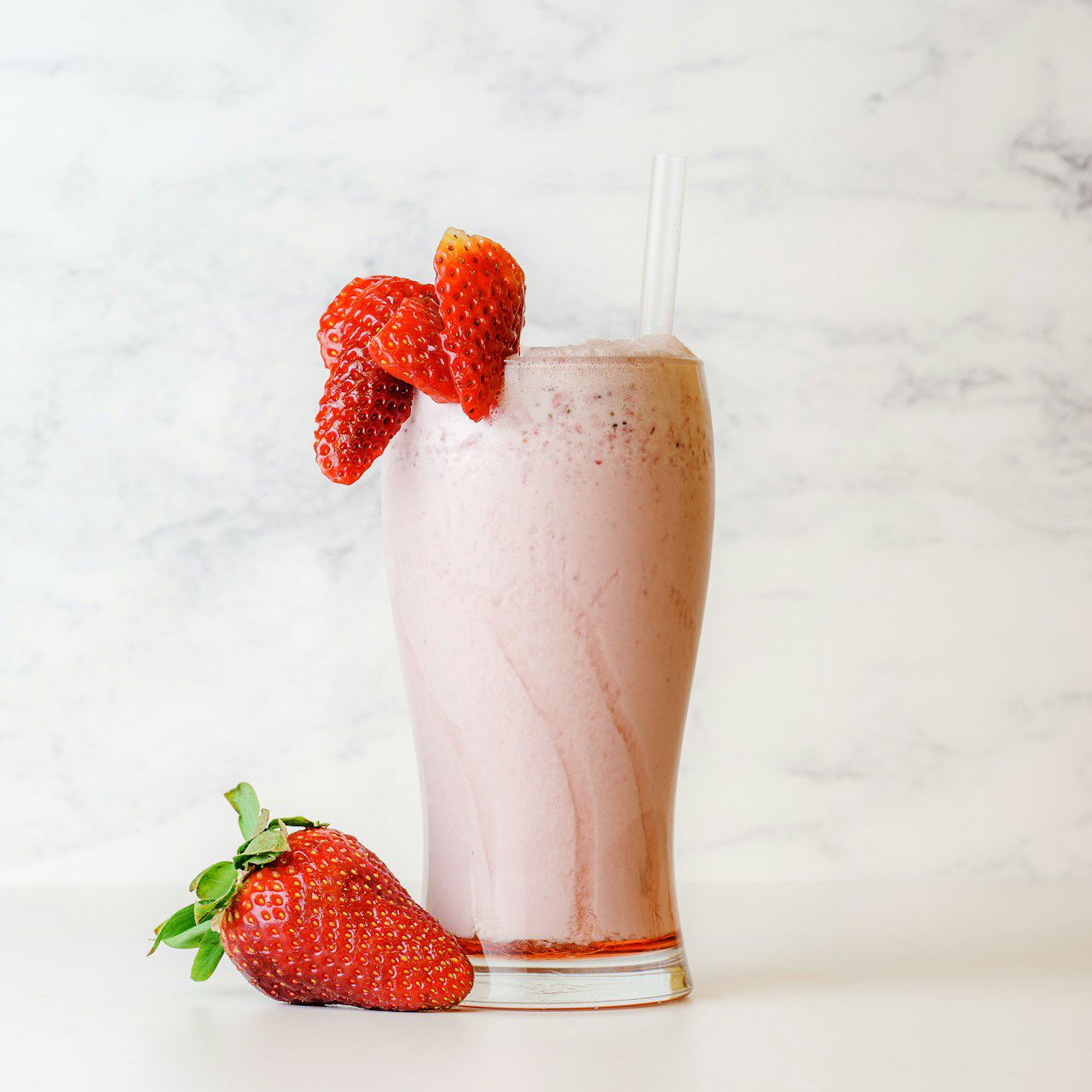 Et højt glas med jordbærmilkshake toppet med to sugerør og pyntet med friske jordbær, placeret på en lys baggrund.