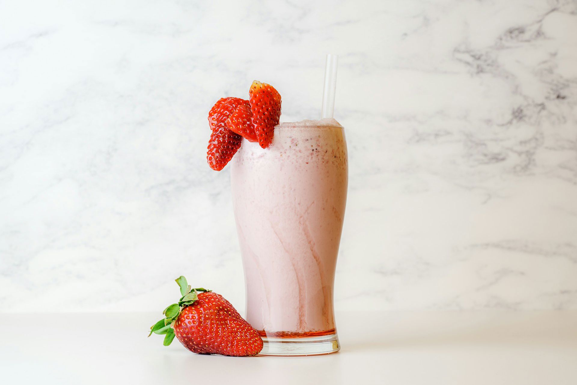 Et højt glas med jordbærmilkshake toppet med to sugerør og pyntet med friske jordbær, placeret på en lys baggrund.