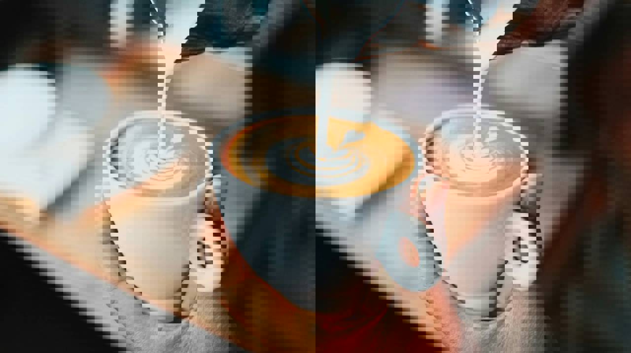 Nærbillede af en hånd, der holder en hvid kop cappuccino, mens mælk hældes i og danner latte art.