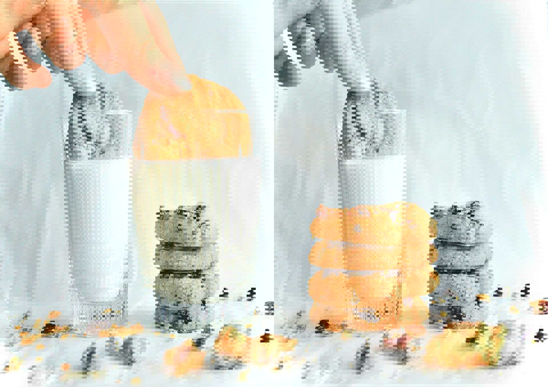 En hånd dypper en småkage i et glas mælk, mens en stak af fire småkager ligger ved siden af på et hvidt underlag med krummer omkring.